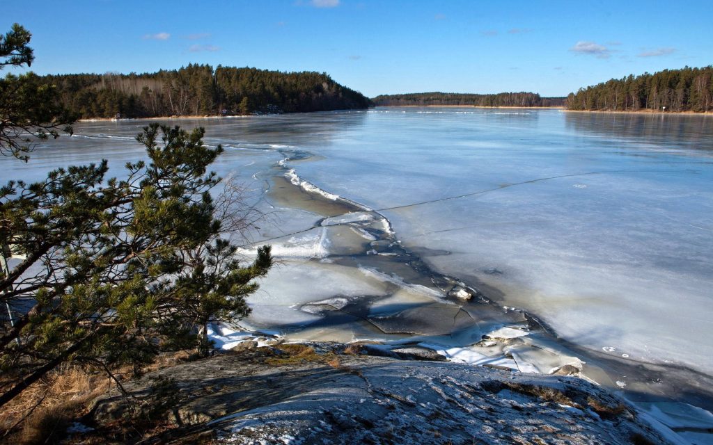 Frozen lake with cracked ice pine tree rocky snow snowy foreground under vivid blue sky winter Wallpaper