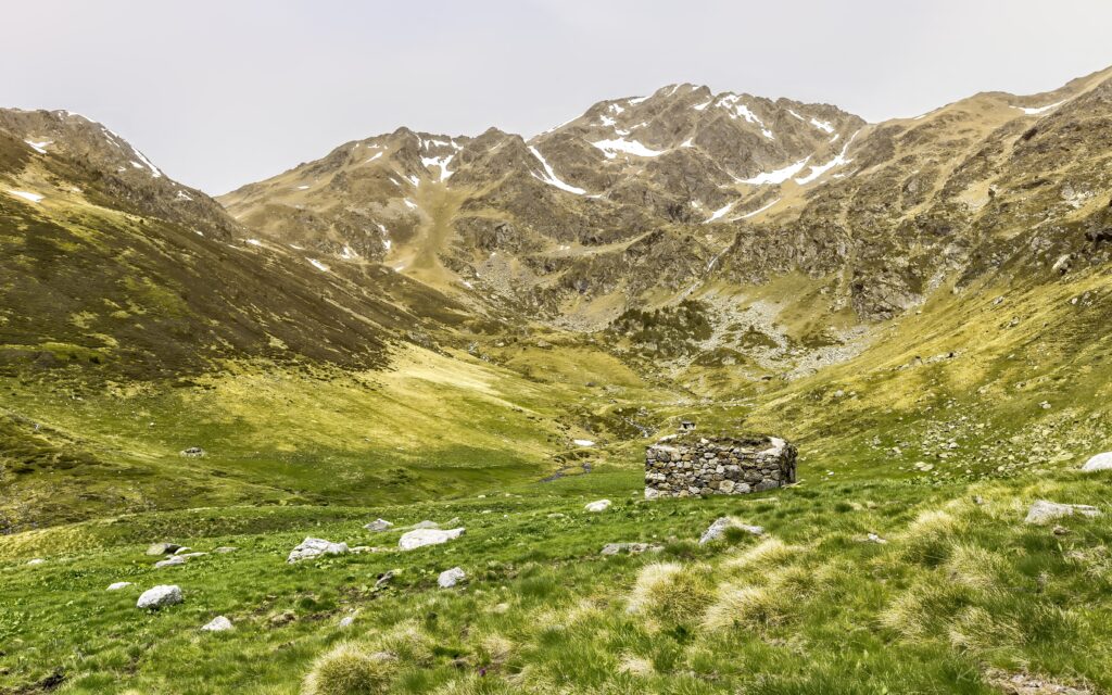 Stone well in lush green valley surrounded by rugged snow-capped mountains under soft overcast sky Wallpaper