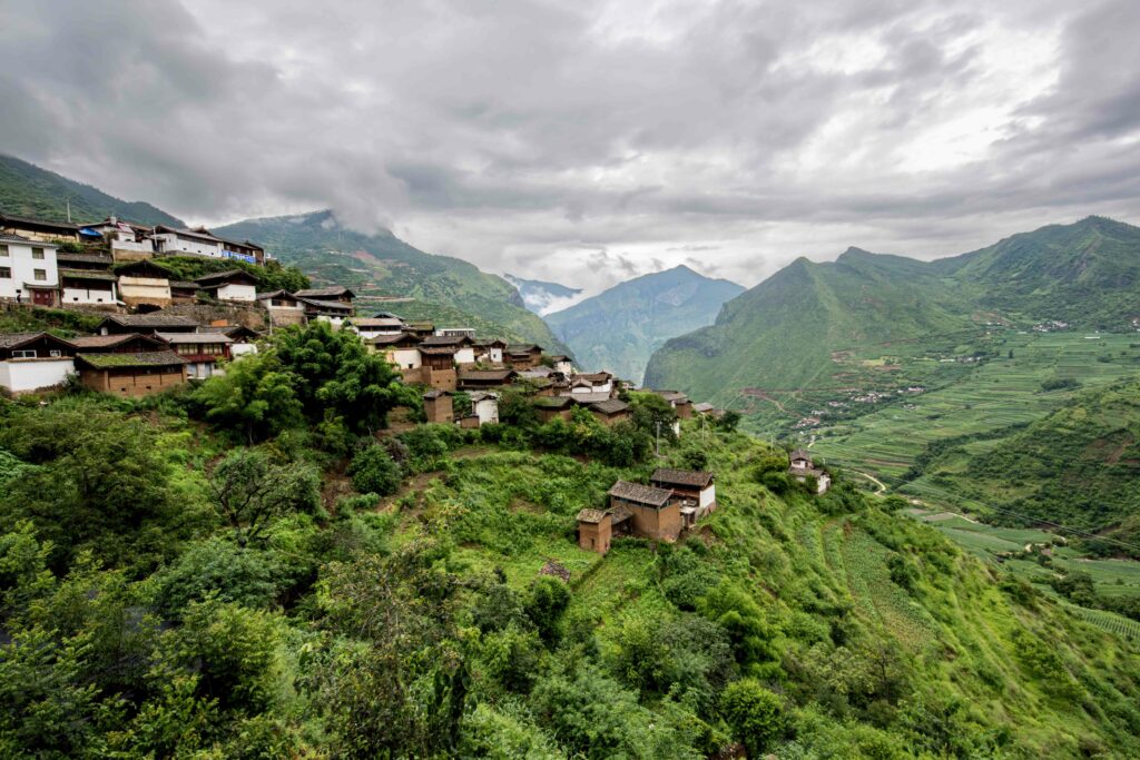 Serene hillside village with rustic houses lush greenery dramatic mountains cloudy sky wallpaper