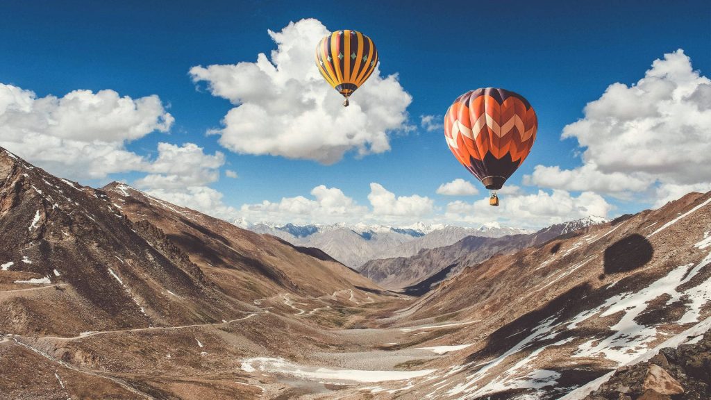 Two colorful hot air balloons float over rugged mountain valley with snow peaks and blue sky Wallpaper