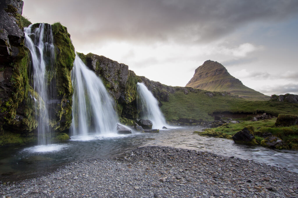 Serene Iceland waterfall over mossy rocks flowing into river with mountain peak under cloudy sky Wallpaper