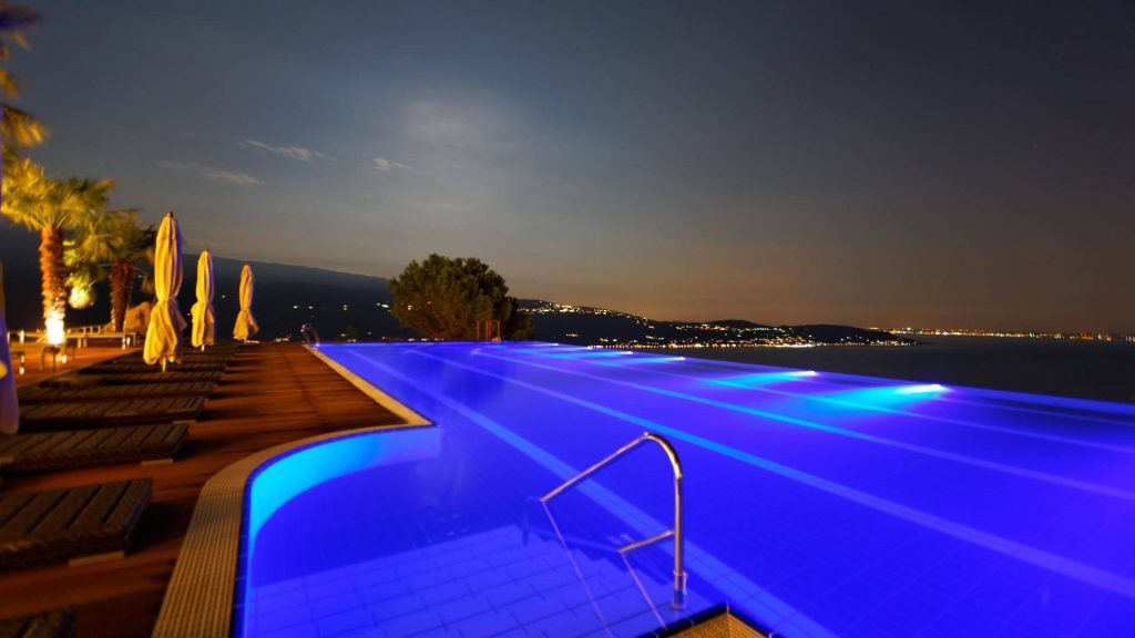 Infinity pool at night with blue lights wooden deck lounge chairs and city skyline view Wallpaper