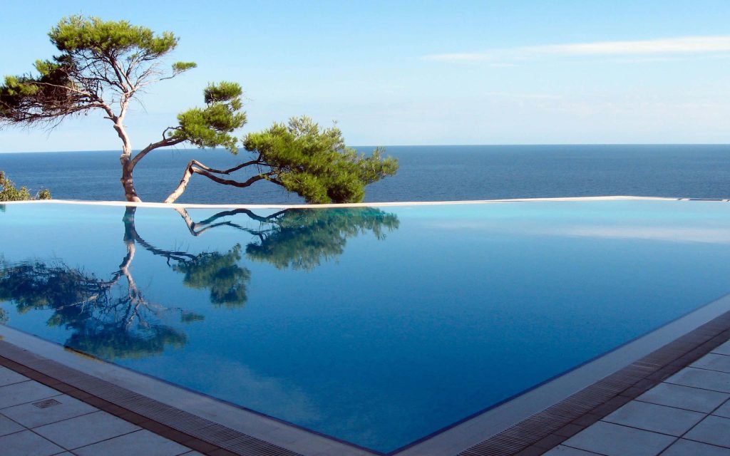 Luxurious infinity pool with leaning pine tree reflection overlooking calm deep blue ocean under sunny sky Wallpaper