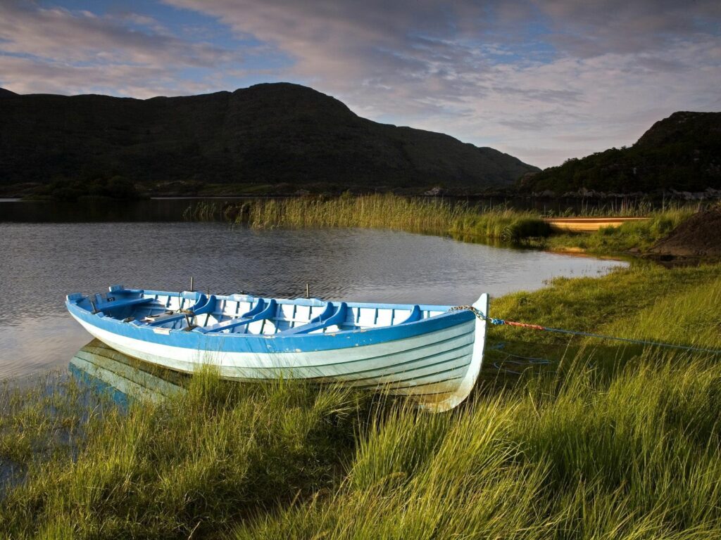 Light blue wooden boat moored on grassy Irish lake shore with calm waters and cloudy sky Wallpaper