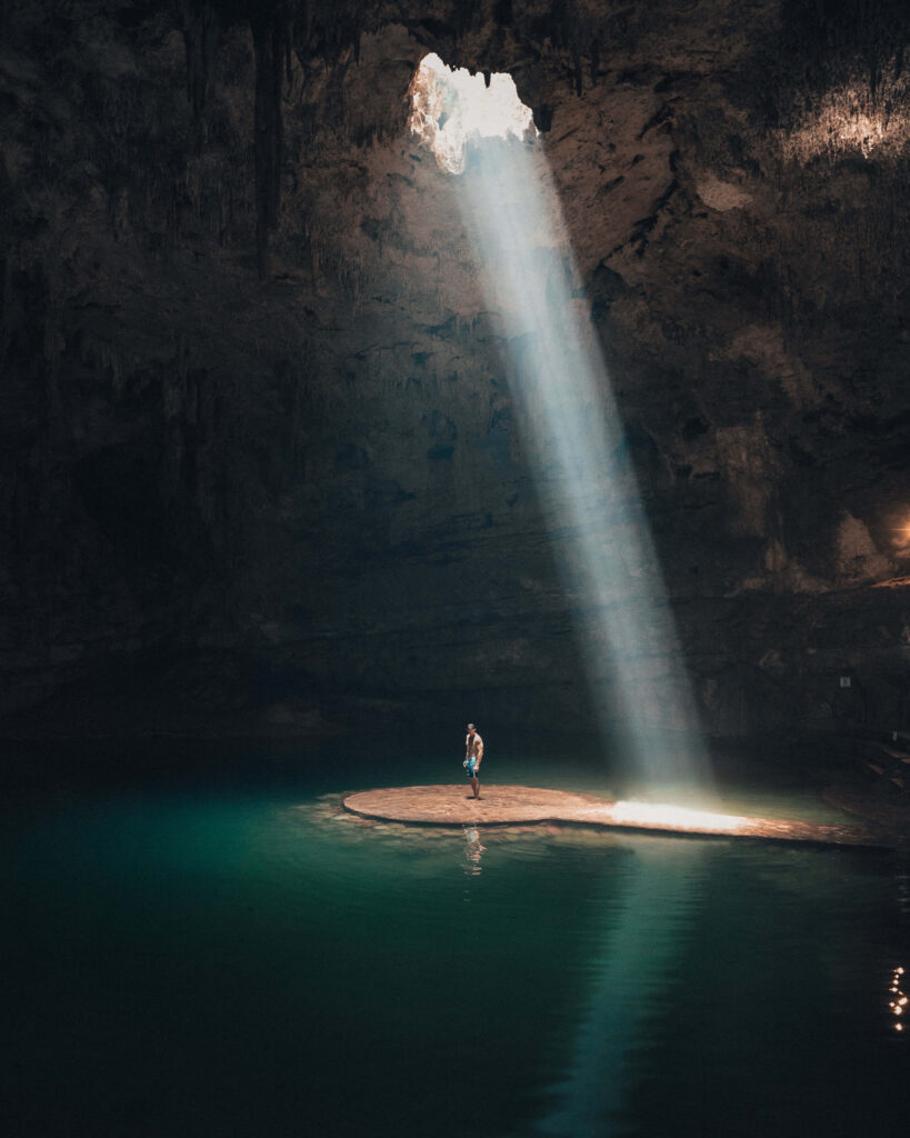 Lone Figure in Cave Island Surrounded by Emerald Water & Sunlight Wallpaper