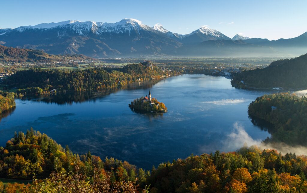Serene island with classic tower on lake surrounded by autumn trees and snow-capped mountains Wallpaper