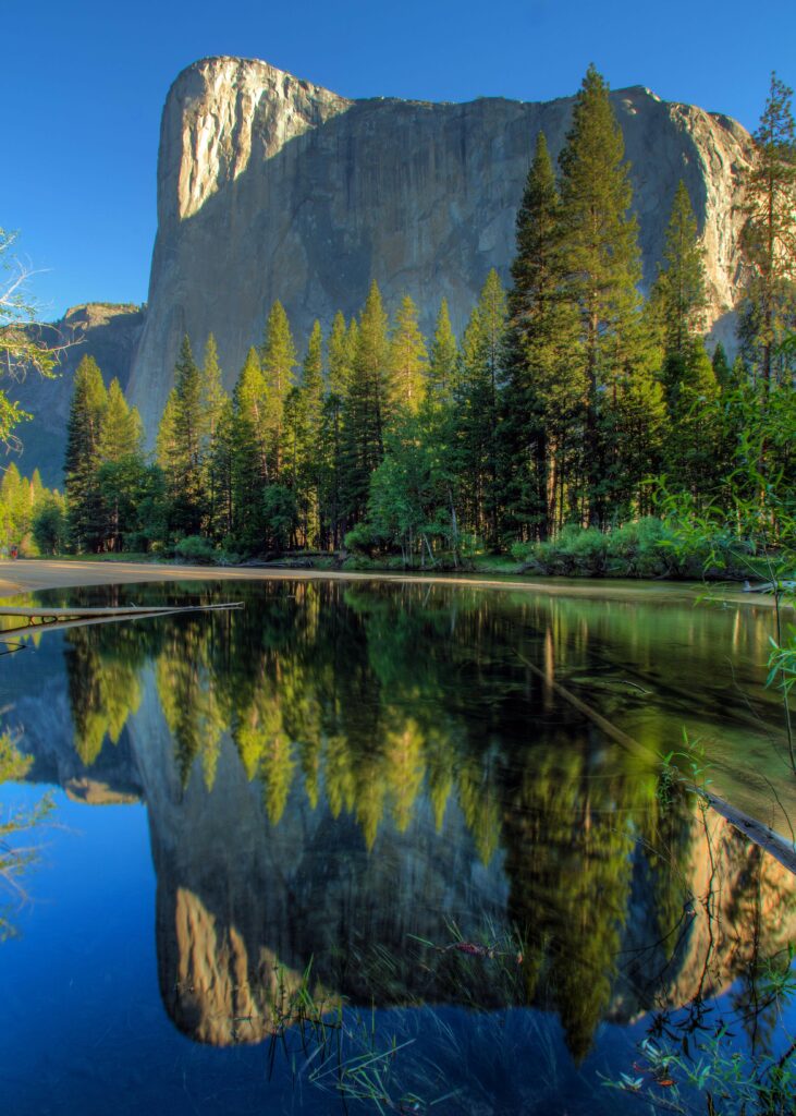 Tranquil lake reflects lush green trees and towering rock under bright blue sky landscape wallpaper