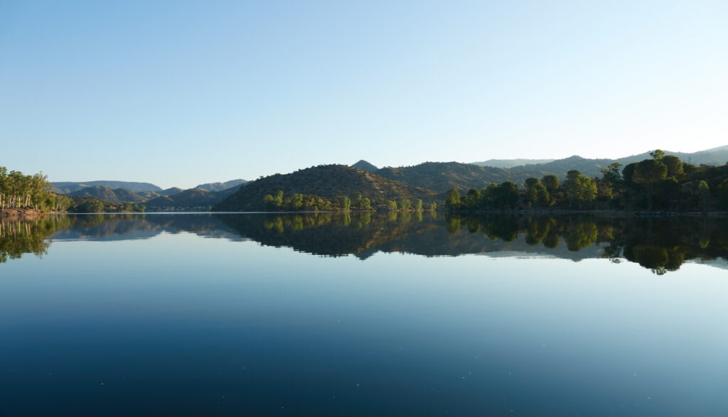 Calm lake reflecting lush forest and distant hills under clear blue sky serene nature wallpaper