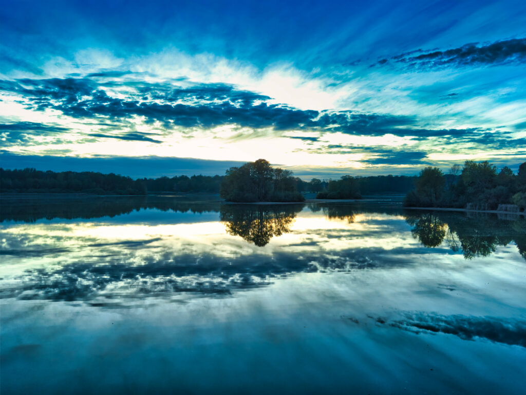 Serene lake with dense forest and dramatic sky reflected on calm water in vivid blue and green Wallpaper