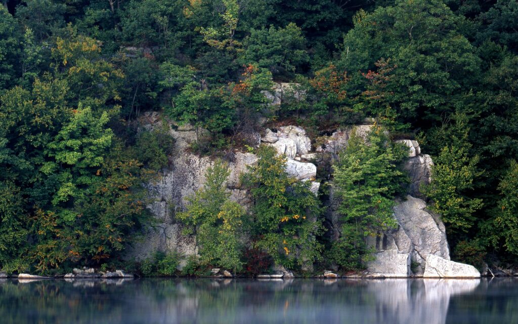Serene lake with rocky shore and lush green trees reflected on smooth calm water background Wallpaper