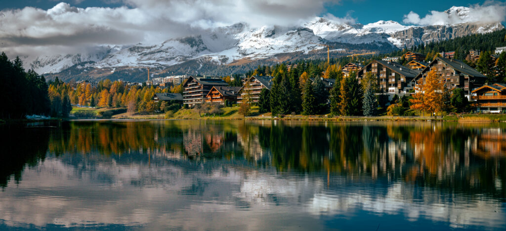 Serene lake reflecting autumn trees charming houses and snow-capped mountains vibrant landscape Wallpaper