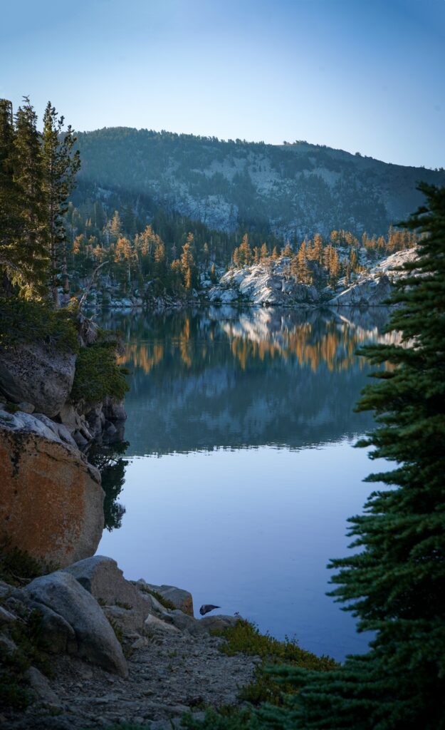 Serene lake with rugged rocks and evergreen trees reflecting forested mountains and bright sky Wallpaper