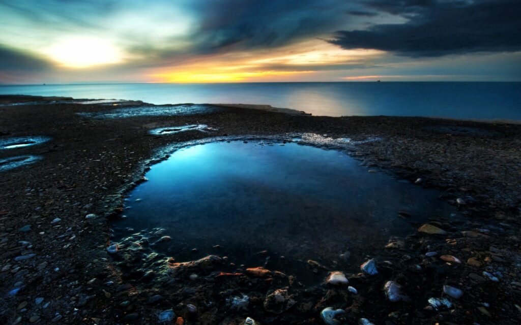 Tranquil reflective lake with dark wet stones under moody overcast sky at sunset wallpaper background