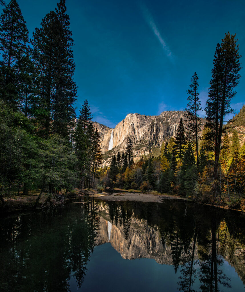 Serene lake reflecting pine trees rocky cliffs and waterfall under vibrant blue sky wallpaper