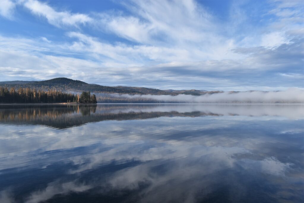 Serene lake reflecting sky clouds pine trees and distant hills with morning mist peaceful landscape Wallpaper
