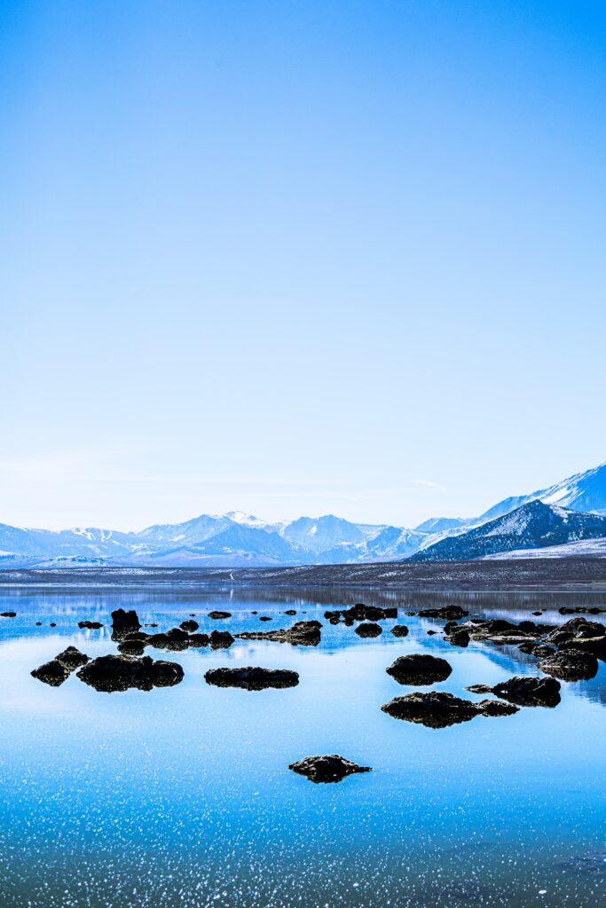 Tranquil lake with clear water reflecting rugged rocks and snow-capped mountains under blue sky Wallpaper