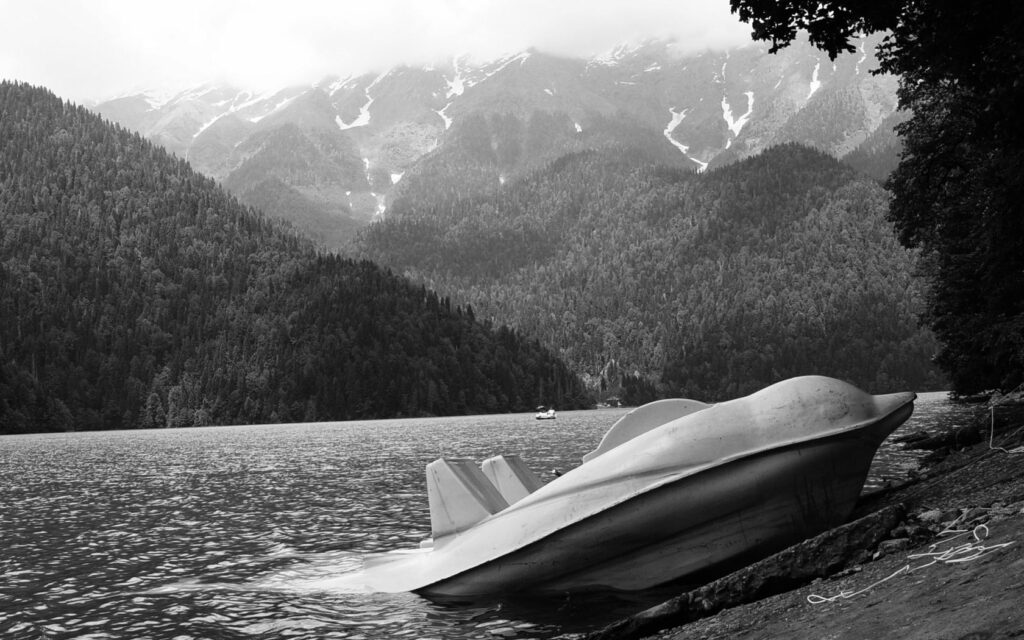 Black and white Lake Ritsa wallpaper catamaran on rocky shore with misty mountains and snowy peaks
