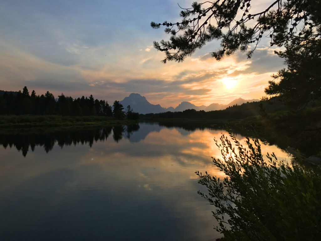 Serene lake at sunset with golden reflections framed by silhouettes lush greenery and distant mountains Wallpaper