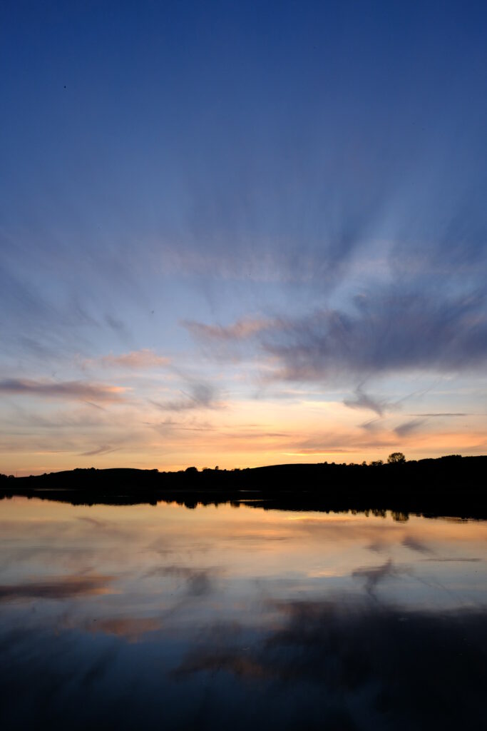 Serene lake at sunset reflecting dramatic sky and wispy clouds with calm water and warm tones Wallpaper