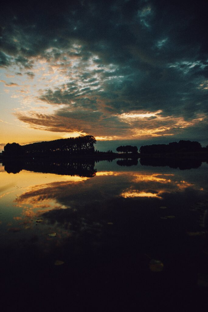 Serene lake at dusk with glowing sunset reflection and silhouetted trees under dramatic clouds Wallpaper