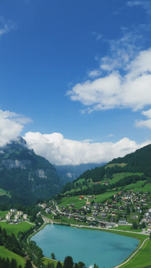 Aerial view of serene lake surrounded by green mountains and a peaceful village under blue sky Wallpaper