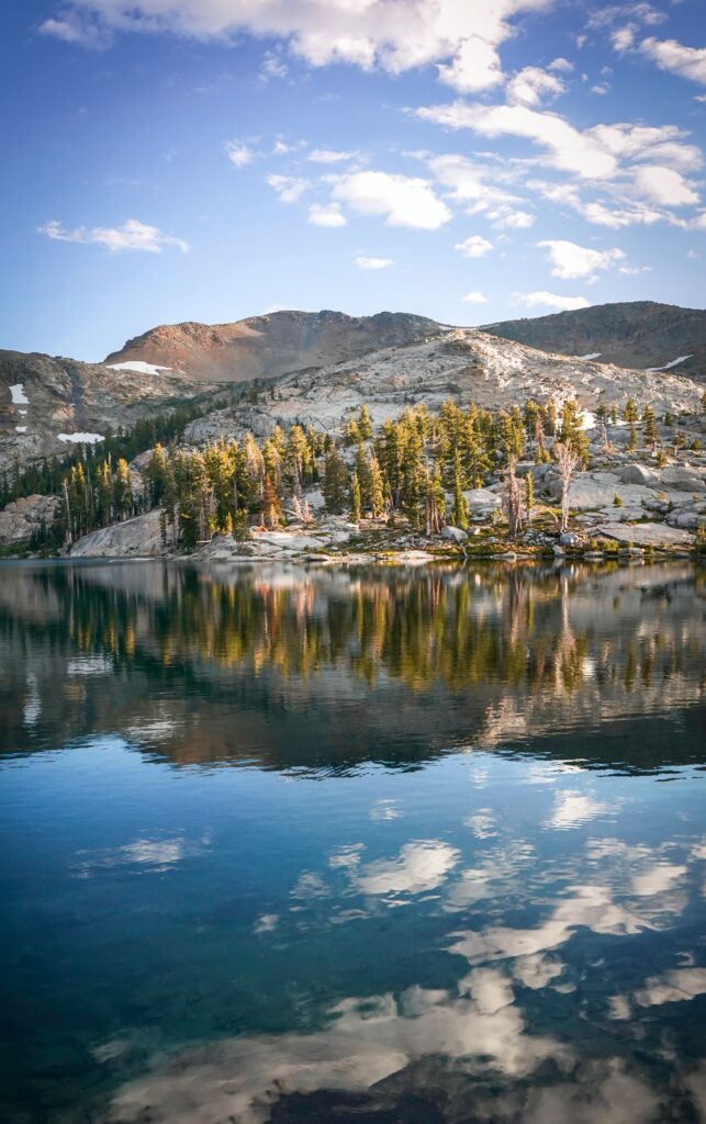 Serene lake with pine trees and rugged rocks reflecting sky clouds in warm natural light wallpaper