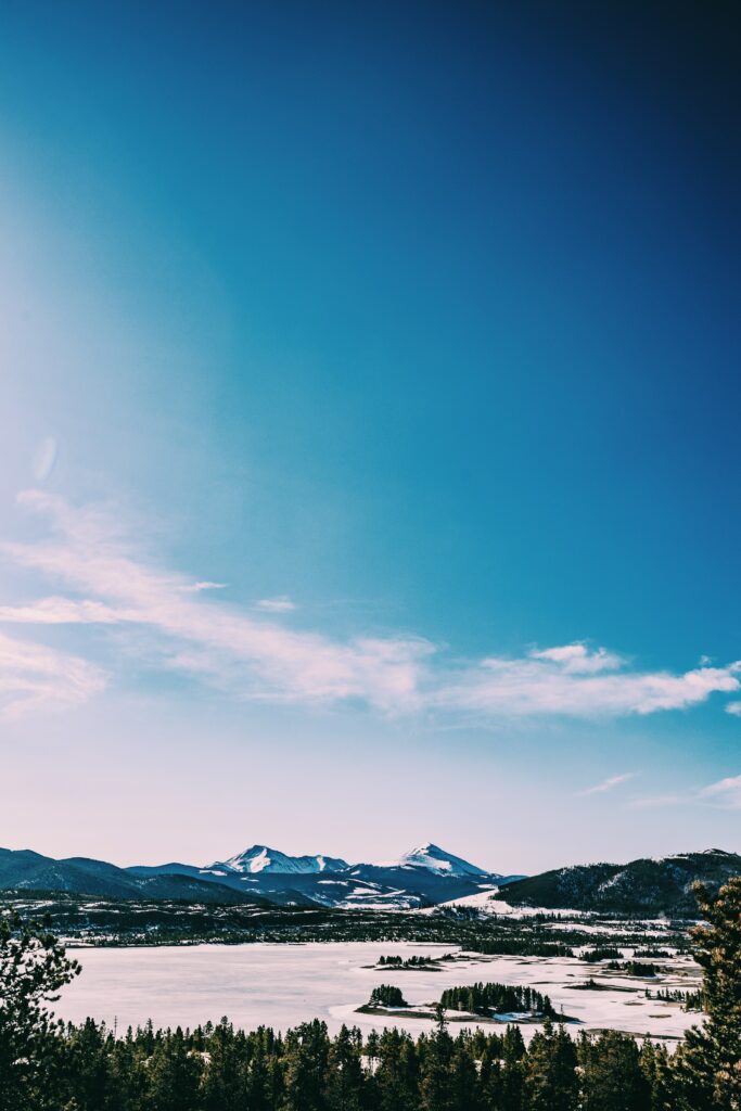 Serene lake with dense forest and snow-capped mountains under a vast blue sky wallpaper background