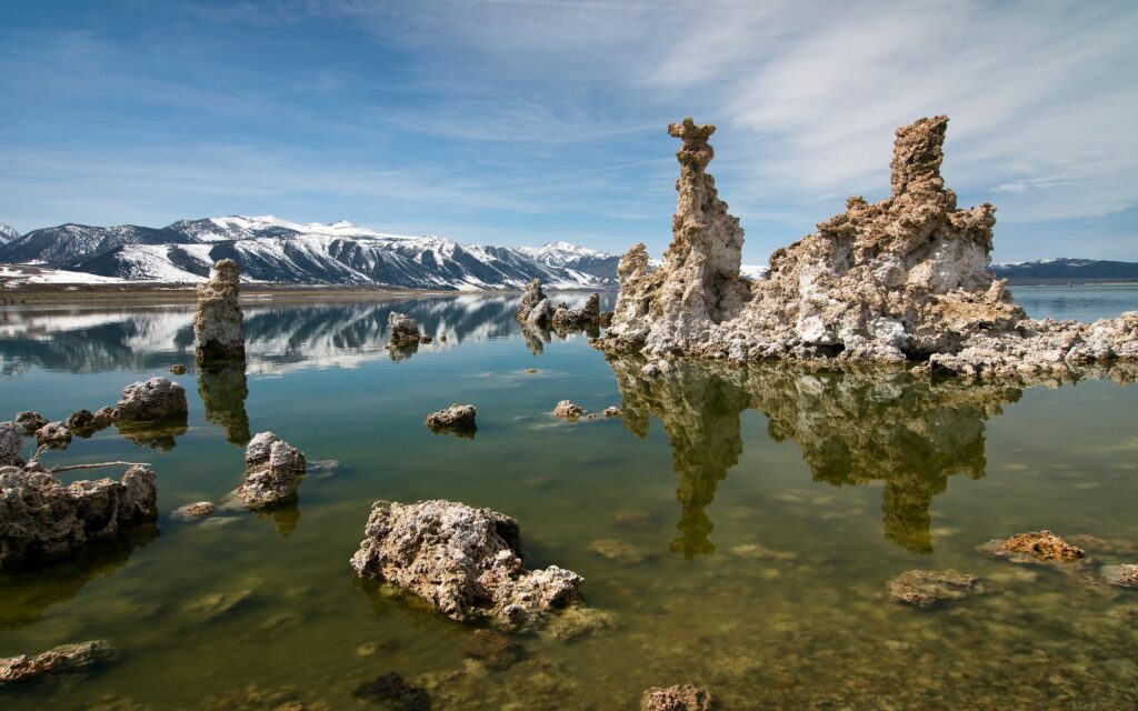 Dramatic rock formations rise from clear lake waters with snowy mountains and calm sky backdrop Wallpaper
