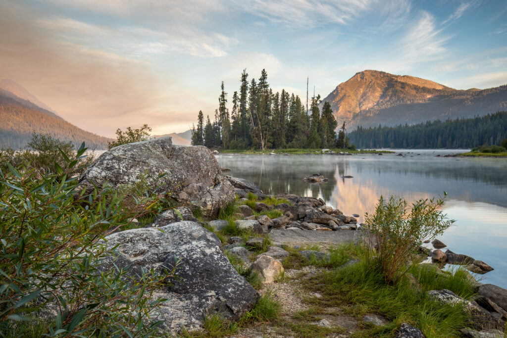 Serene lakeside with green trees rugged rocks and mountains reflecting in calm water wallpaper background
