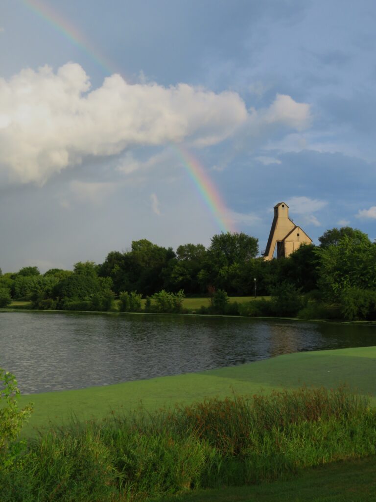 Serene lake with lush green trees and a rainbow over unique architectural building background Wallpaper