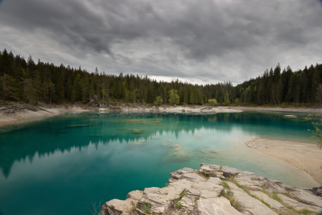 Serene turquoise lake reflecting pine forest under cloudy sky with rocky shoreline nature wallpaper
