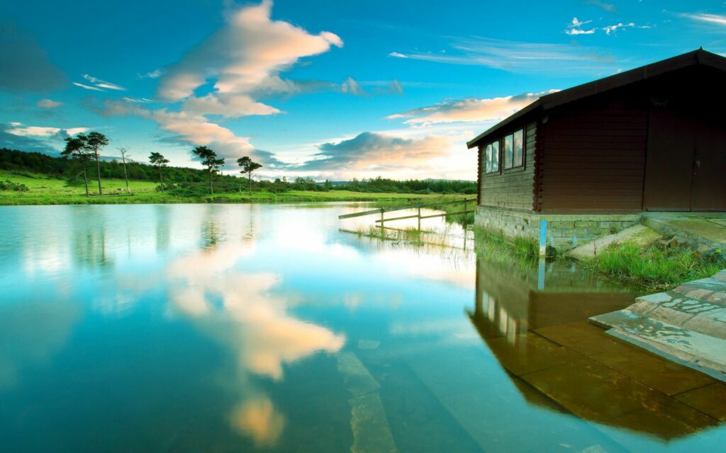 Peaceful dark wooden lodge by tranquil lake with vivid blue sky and mirror-like water reflection Wallpaper