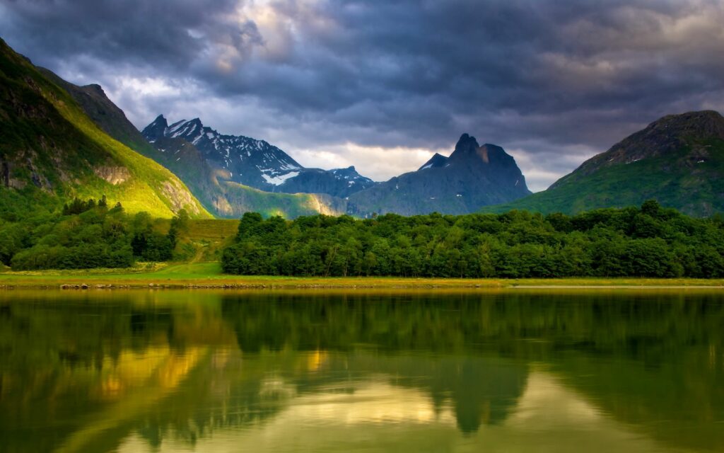 Serene lakeside with dark clouds over mountains lush trees reflected on calm water golden light Wallpaper