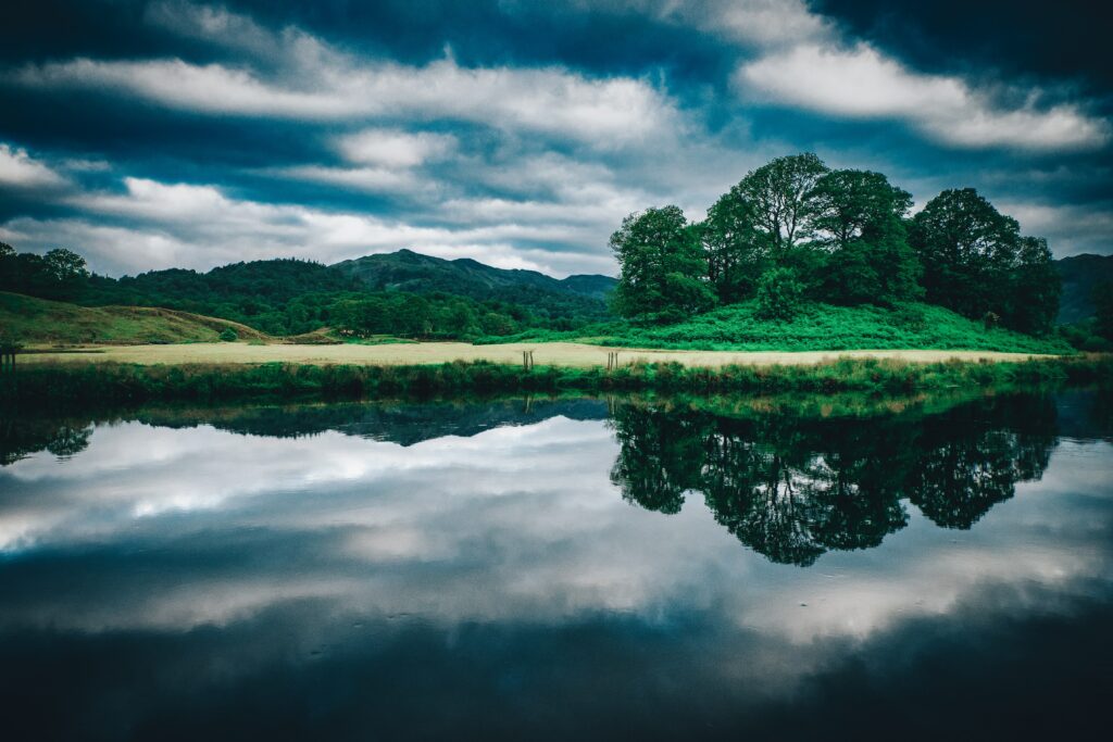 Serene lakeside wallpaper with lush green trees and dark clouds reflecting on calm water surface