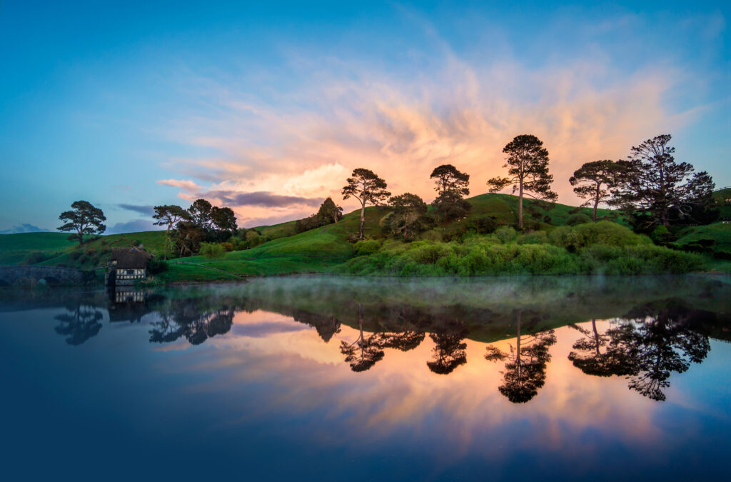 Serene lakeside sunset with green hills and tree silhouettes reflected on calm water wallpaper background
