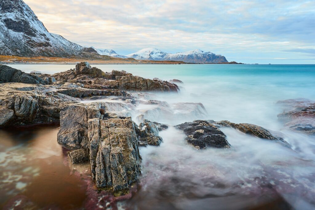Serene lakeside with rugged rocks turquoise water and snow-capped mountains under cloudy sky Wallpaper