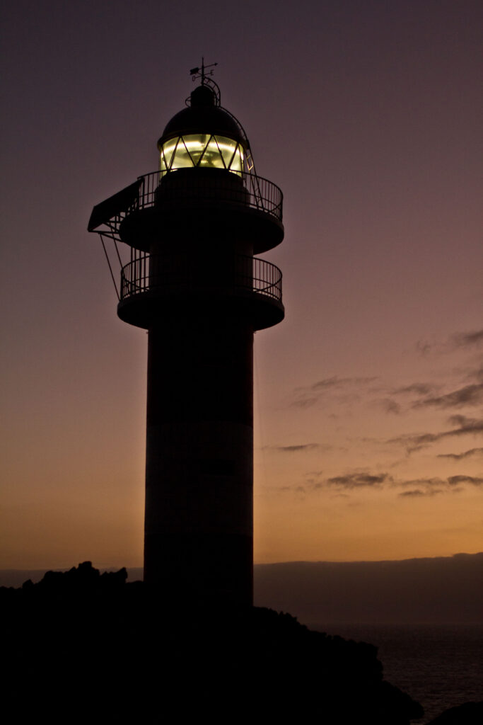Silhouette of lighthouse glowing on rocky coast at sunset with warm purple and orange sky Wallpaper