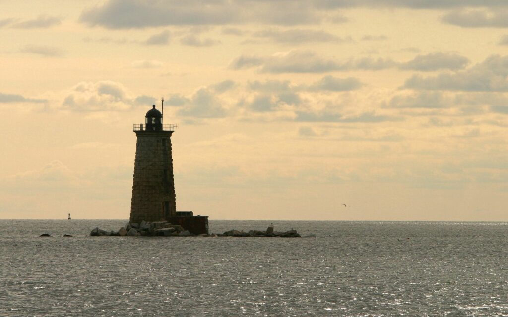 Lighthouse silhouette at sunset over calm sea with glowing light patches and soft clouds wallpaper
