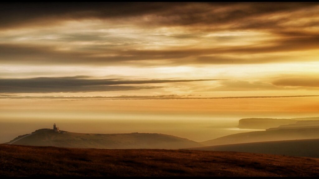Serene lighthouse on rugged foggy coastline with golden sunlight and dramatic cloudy sky wallpaper
