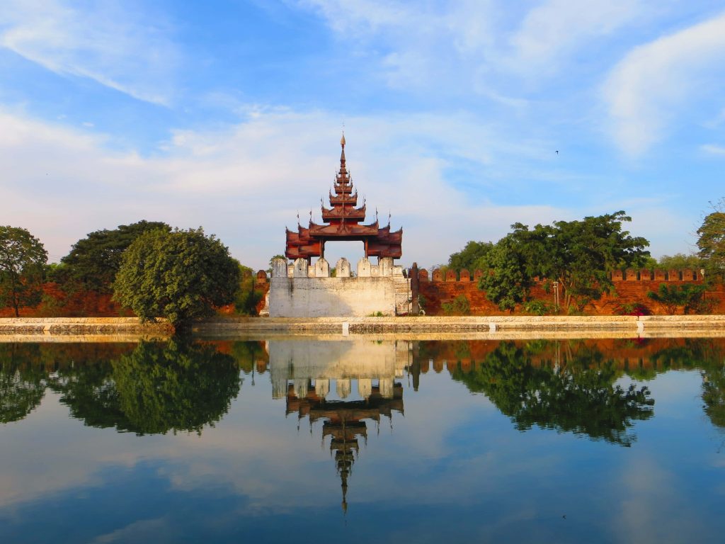 Historic Mandalay Palace with tiered wooden pavilion reflected in calm moat under blue sky Wallpaper