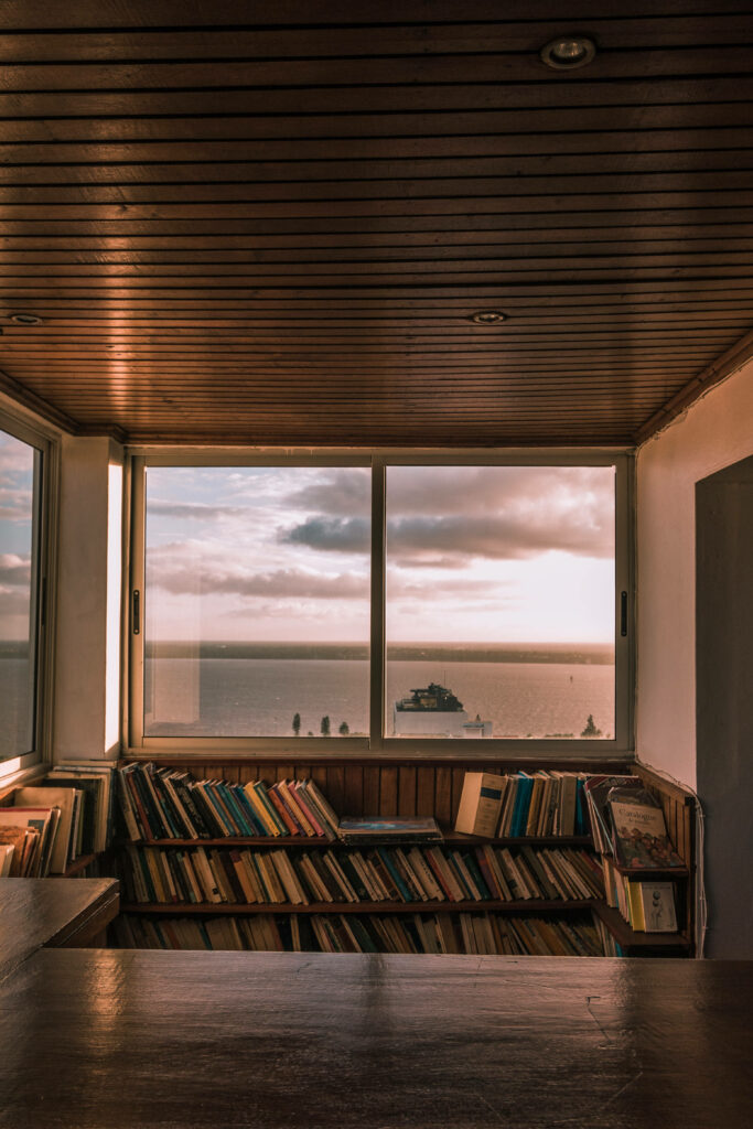 Minimalist library nook with books by large window overlooking sea boat sunset serene view wallpaper