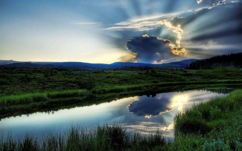 Serene pond in green meadows with sunbeams breaking through clouds in peaceful nature landscape Wallpaper