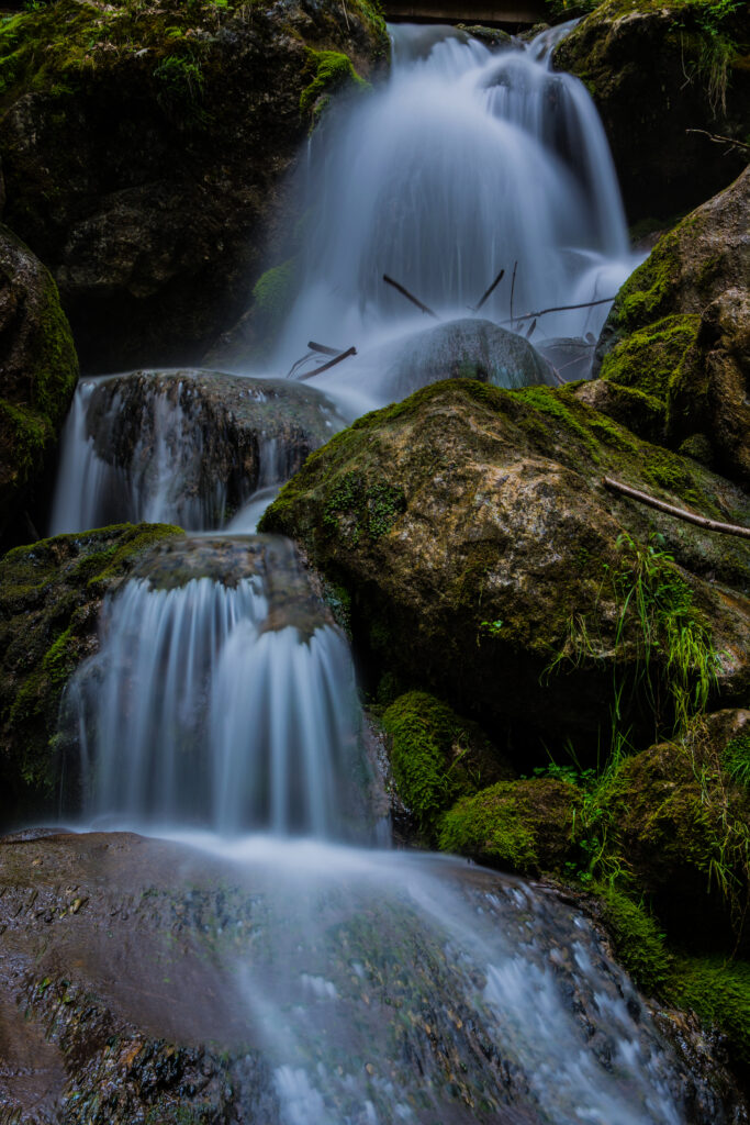 Serene waterfall flowing over mossy stones with lush green foliage and smooth water flow background Wallpaper
