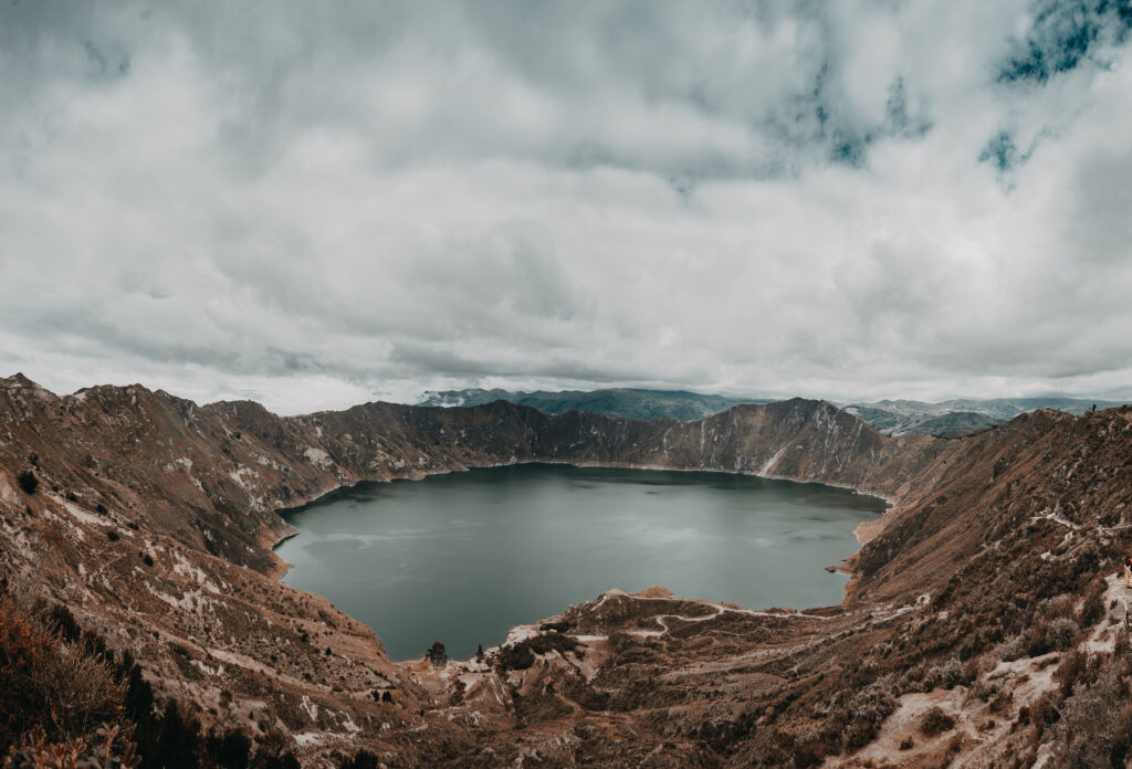 Round mountain lake surrounded by rugged brown and green hills under dramatic cloudy sky Wallpaper