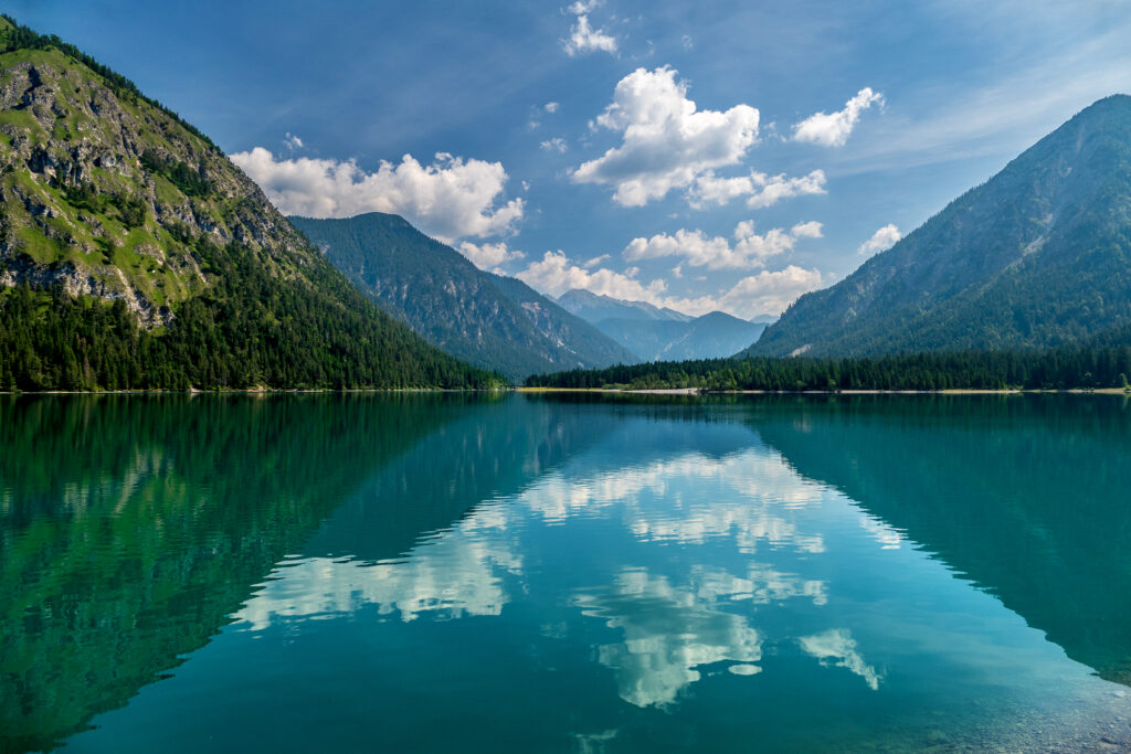 Serene lake reflecting mountains and green slopes under clear blue sky with clouds wallpaper