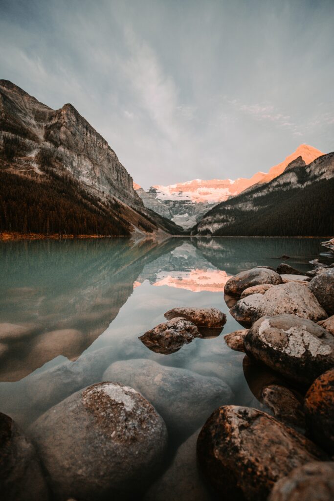 Serene mountain lake with clear water reflecting rugged peaks pastel sky and smooth stones foreground Wallpaper