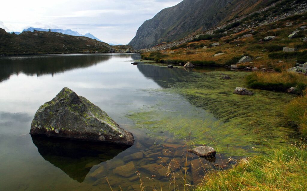 Serene mountain lake reflecting rugged peaks cloudy sky with stones and green algae beneath water Wallpaper