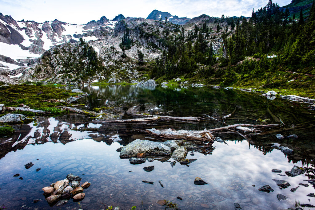 Serene mountain lake reflecting rugged peaks lush green trees and scattered stones vibrant nature landscape Wallpaper