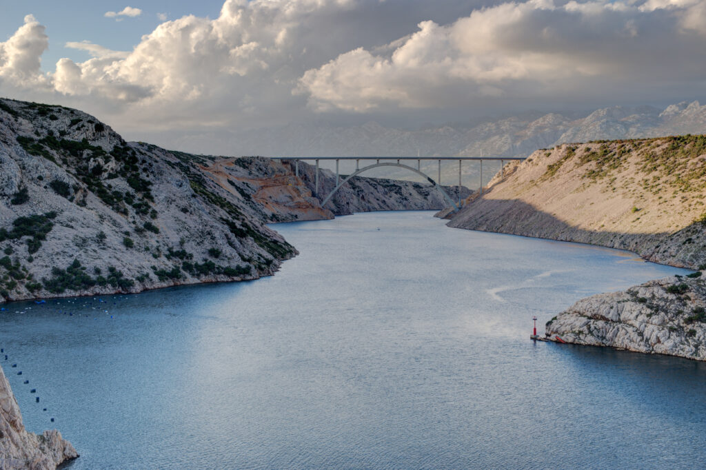 Serene lake winding through rugged cliffs with a graceful bridge and towering mountains background Wallpaper