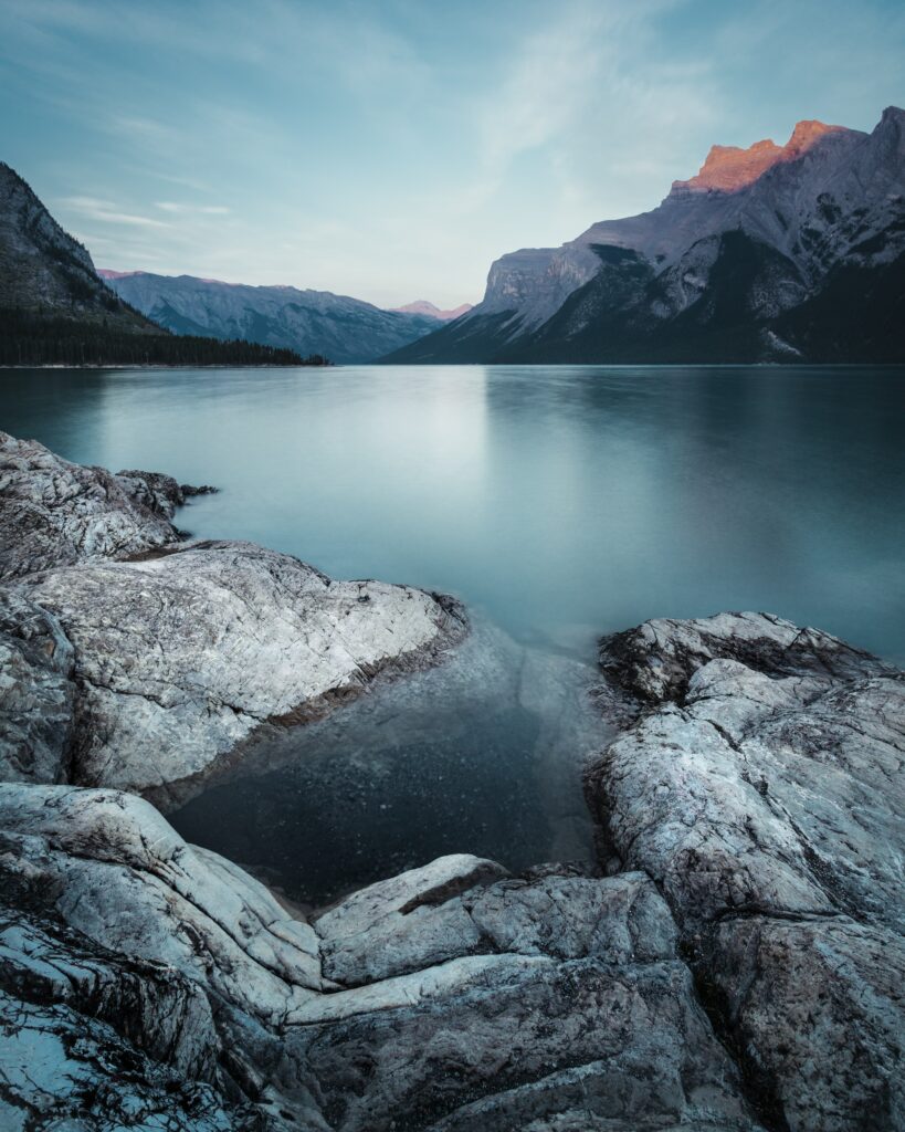 Rugged stones path to calm lake framed by majestic mountains under soft pale blue sky wallpaper